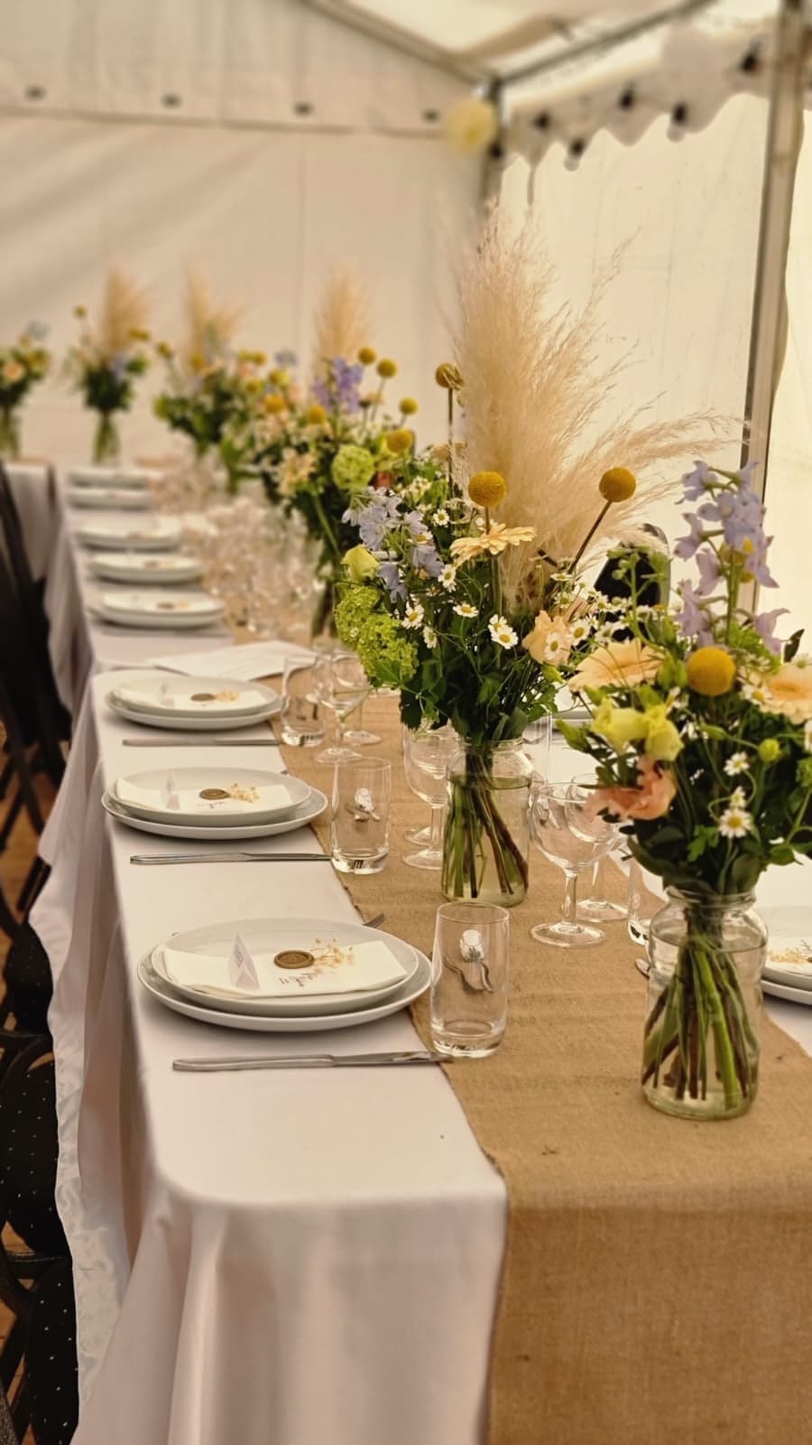 Langbord i festtelt med markblomster, pampasgræs og craspedia i glasvaser